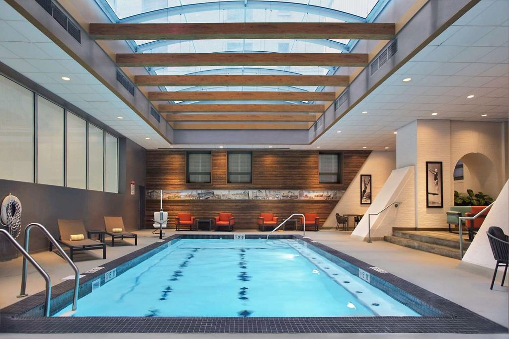 Indoor hotel swimming pool with skylight ceiling, lounge seating, and shared wellness facilities